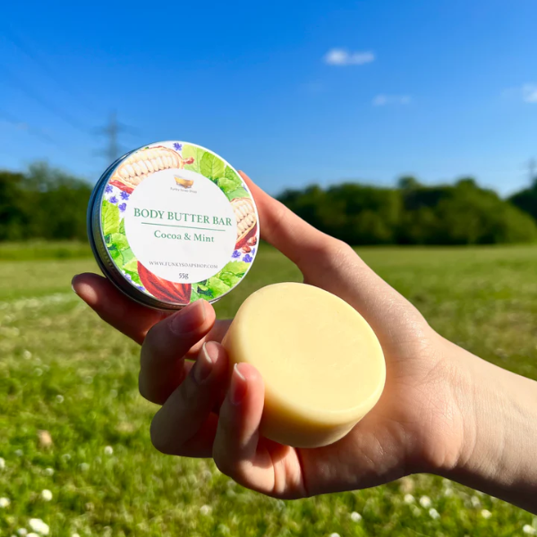 Natural vegan body butter bar in cocoa and mint, shown in aluminium tin with lid of, in the palm of a hand out in the countryside