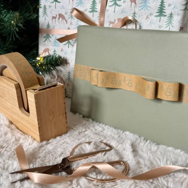 Christmas paper tape brown with green Christmas trees shown on roll in bamboo dispenser next to a parcel wrapped in green paper with the tape making a decorative shape