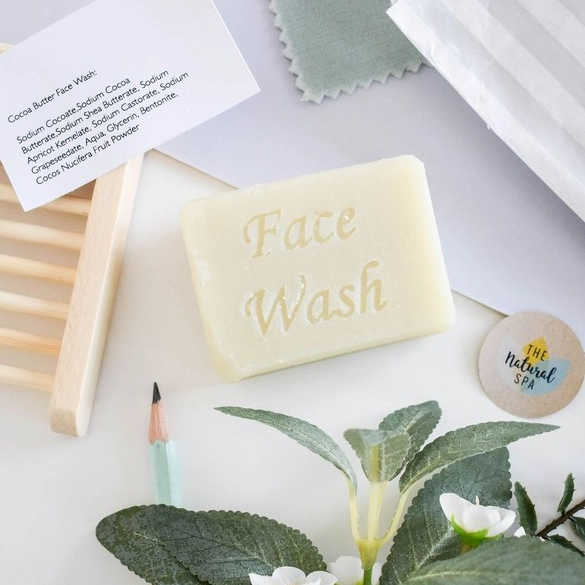 Travel facial soap bar (cocoa butter) shown with greenery and a wooden soapdish, the words 'Facial wash' stamped on the bar