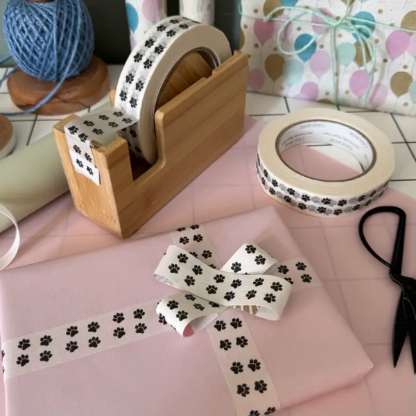 Roll of white paper tape with black pawprint design shown on the roll in a bamboo dispenser and applied to a wrapped gift in a bow shape