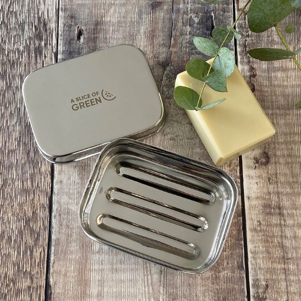 Travel soap or snack tin with lid shown off to see ridged base inside with a bar of soap laying alongside. Lid has logo reading A Slice of Green.