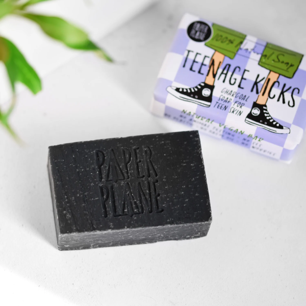 Teenage kicks soap bar shown unwrapped (a black charcoal soap bar) next to a wrapped bar in paper packaging with a cartoon image of a teenager's legs in trainers