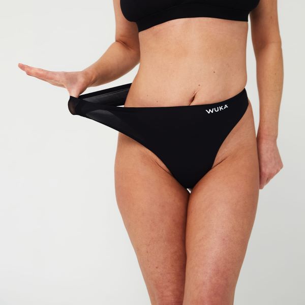 Black stretch thong period pants shown on model from front, with a hand pulling the pants to show the stretch