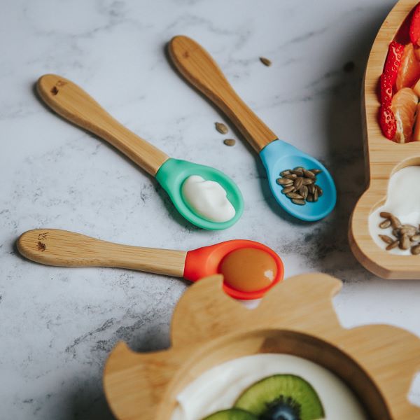 Bamboo and silicone baby weaning spoons in blue, green and orange with some puree and seeds on each, shown alongside bamboo bowls  with food inside