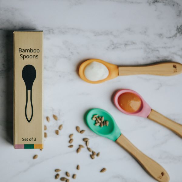 Bamboo and silicone baby weaning spoons in pink, green and yellow, with some puree and seeds on each, shown alongside cardboard packaging