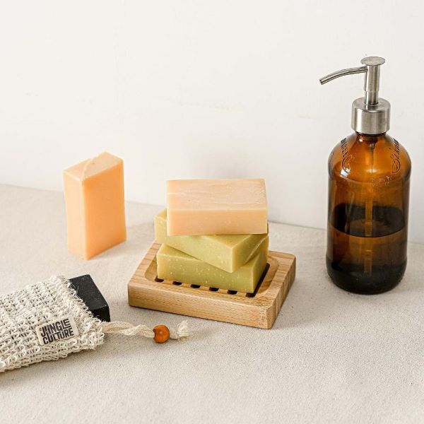 Bamboo soapdish shown with bars of soap next to a glass bottle of soap with pump dispenser and a sisal soap bag  