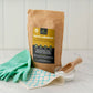 Borax substitute in paper bag alongside a wooden scoop, compostable cloth and latex gloves