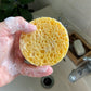 Cellulose facial sponge, round, shown in a soapy hand next to a sink