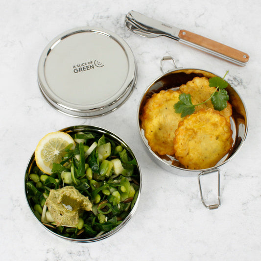 Stainless steel stackable round lunch boxes with food on a white surface, featuring the brand 'A Slice of Green', alongside cutlery.