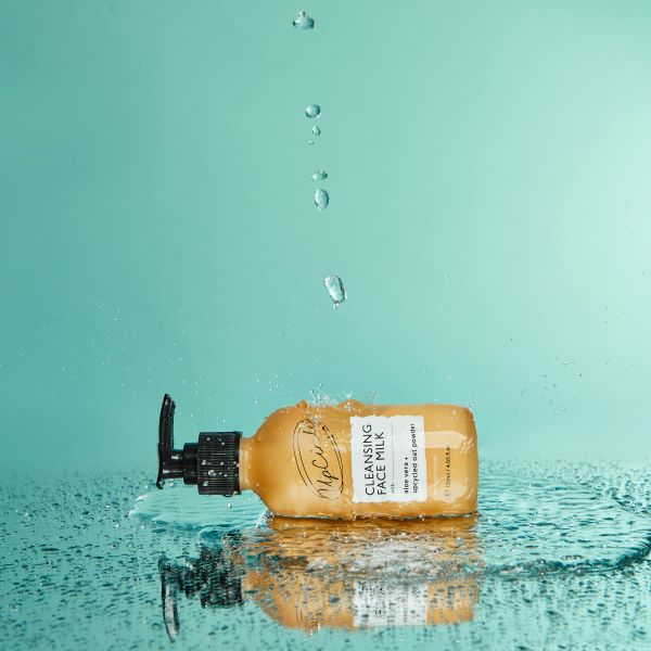 Cleansing face milk in glass bottle with black pump, showing on its side with water pouring down and aqua background