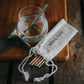 Steel cocktail straw set in rainbow (multicoloured straws) alongside cotton pouch, with one straw inside a cocktail glass