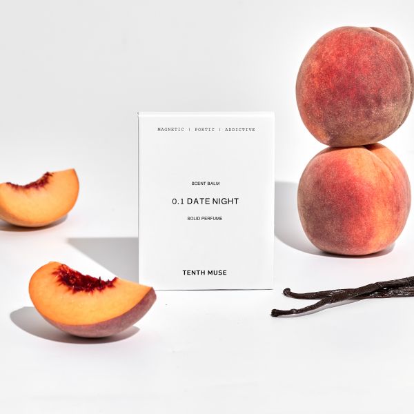 Perfume balm in white cardboard packaging with packaging reading "Magnetic/Poetic/Addictive Date Girl solid perfume" shown alongside peaches cut in half