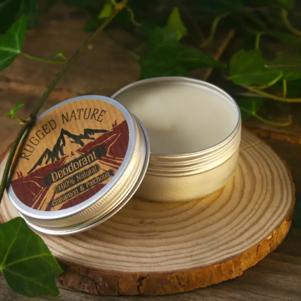 Rugged Nature natural deodorant in a recyclable tin with a label reading "Deodorant 100% natural, cinnamon & patchouli" on a piece of wood with some greenery, tin is open with the deodorant inside visible