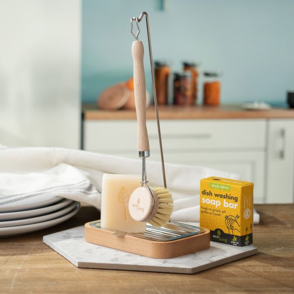 Dish brush holder shown with wooden brush hanging from the hook, and dish soap bar alongside, on a kitchen work surface with dishes close by