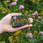Herb garden rosemary soap bar shown wrapped (with text reading "for a moment of quiet calm") sitting in the palm of a hand alongside some wild flowers