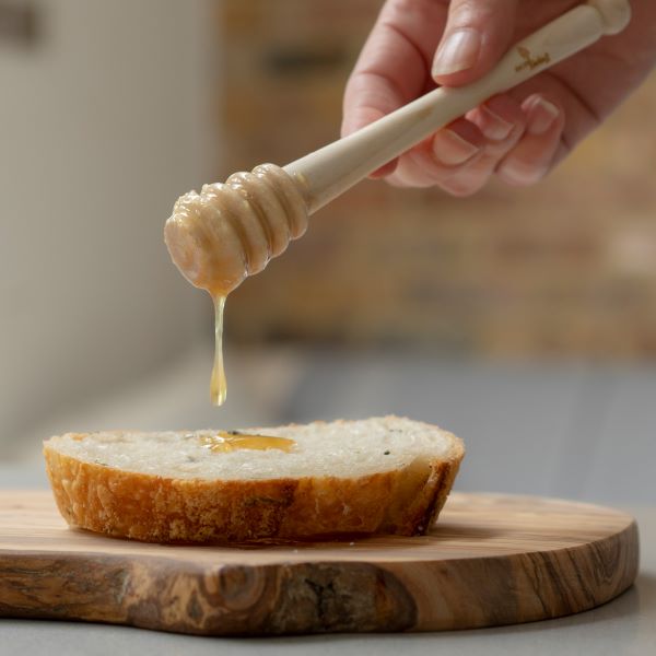 Wooden honey dipper in a hand dribbling honey onto bread sitting on a wooden chopping board