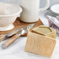 Compostable kitchen scrubber sponge showing the scrubby side of the sponge, next to some dishes and Marseille soap