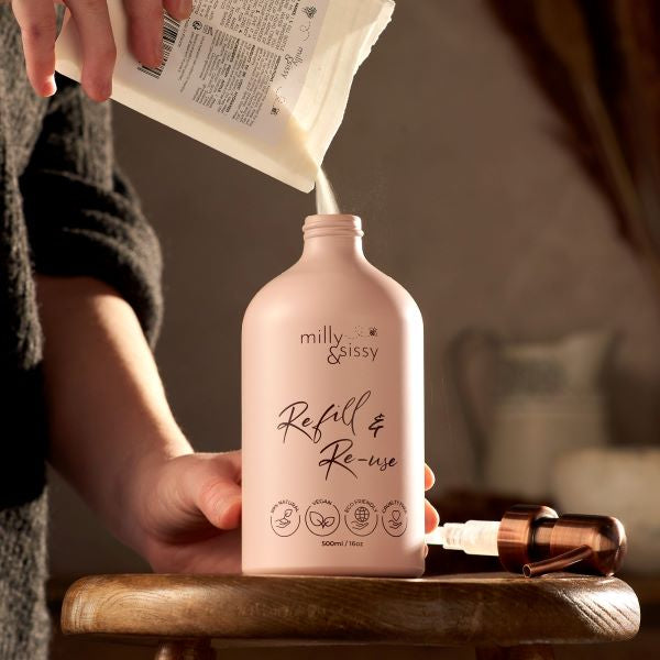 Refillable aluminium bottle in blush pink sitting on a wooden stool with a Milly & Sissy pouch refill being poured in