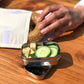 Mini stainless steel mini snack container shown containing cheese and cucumber with a hand lifting a bagel