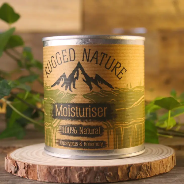 Rugged Nature natural moisturiser in recyclable tin with label reading "Moisturiser 100% natural, eucalyptus and rosemary" sitting on a piece of wood with some greenery