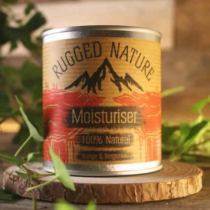 Rugged Nature natural moisturiser in recyclable tin with label reading "Moisturiser 100% natural, orange and bergamot" sitting on a piece of wood with some greenery 