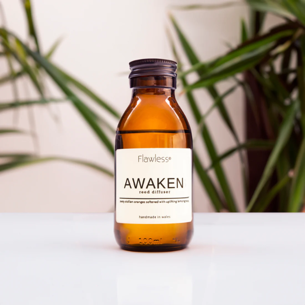 Eco reed diffuser home fragrance amber glass bottle with label reading "Awaken reed diffuser", sitting on a table with plants behind