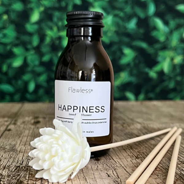 Eco reed diffuser home fragrance amber glass bottle with label reading "Happiness reed diffuser", sitting on a table outside with plants behind, reeds lying alongside