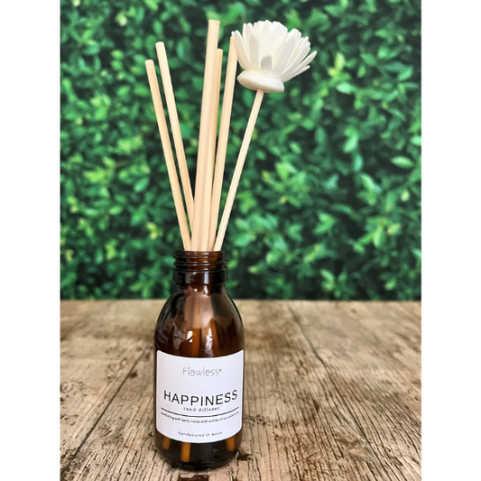 Eco reed diffuser home fragrance amber glass bottle with reeds coming out, with label reading "Happiness reed diffuser", sitting on a table outside with plants behind