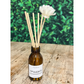 Eco reed diffuser home fragrance amber glass bottle with reeds coming out, with label reading "Opulence reed diffuser", sitting on a table outside with plants behind