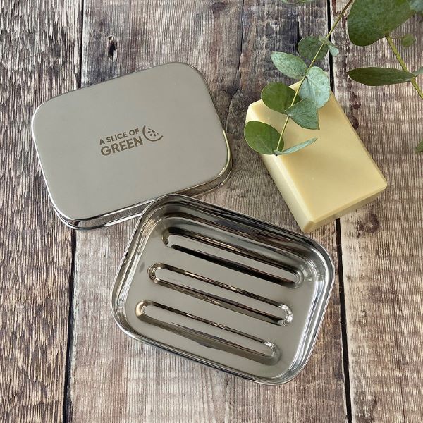 Travel soap or snack tin with lid shown off to see ridged base inside with a bar of soap laying alongside. Lid has logo reading A Slice of Green. 