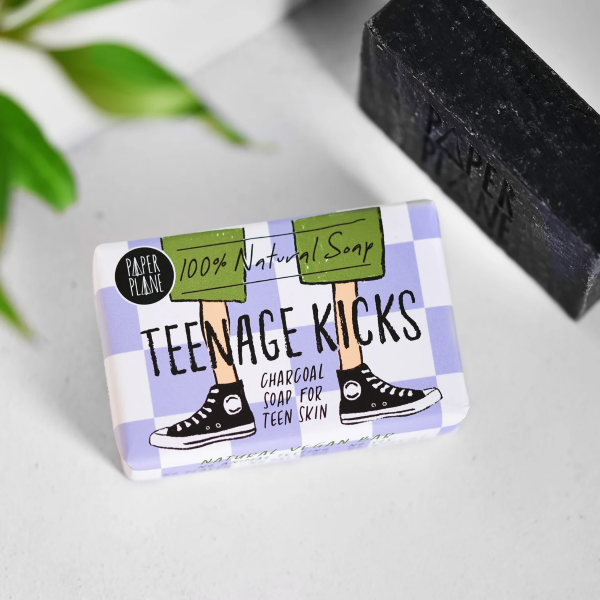 Teenage kicks soap bar shown unwrapped (a black charcoal soap bar) next to a wrapped bar in paper packaging with a cartoon image of a teenager's legs in trainers 