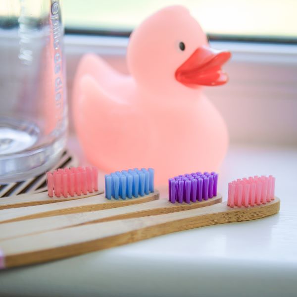 Kids bamboo toothbrush set, pack of 4 in candy colourways (pinks and purples) shown alongside a pink rubber duck in a bathroom