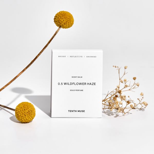 Perfume balm in white cardboard packaging with packaging reading "Bright/Reflective/Grounded Wildflower Haze solid perfume" shown alongside dried flowers