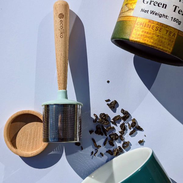 Tea strainer with wooden handle, alongside a can of green tea leaves on a light blue background.
