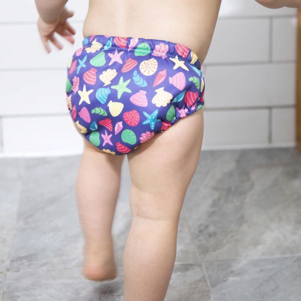 Baby standing wearing reusable swim pants back view in Mussel Seashell design (navy blue background with colourful starfish, mussels and shells) 