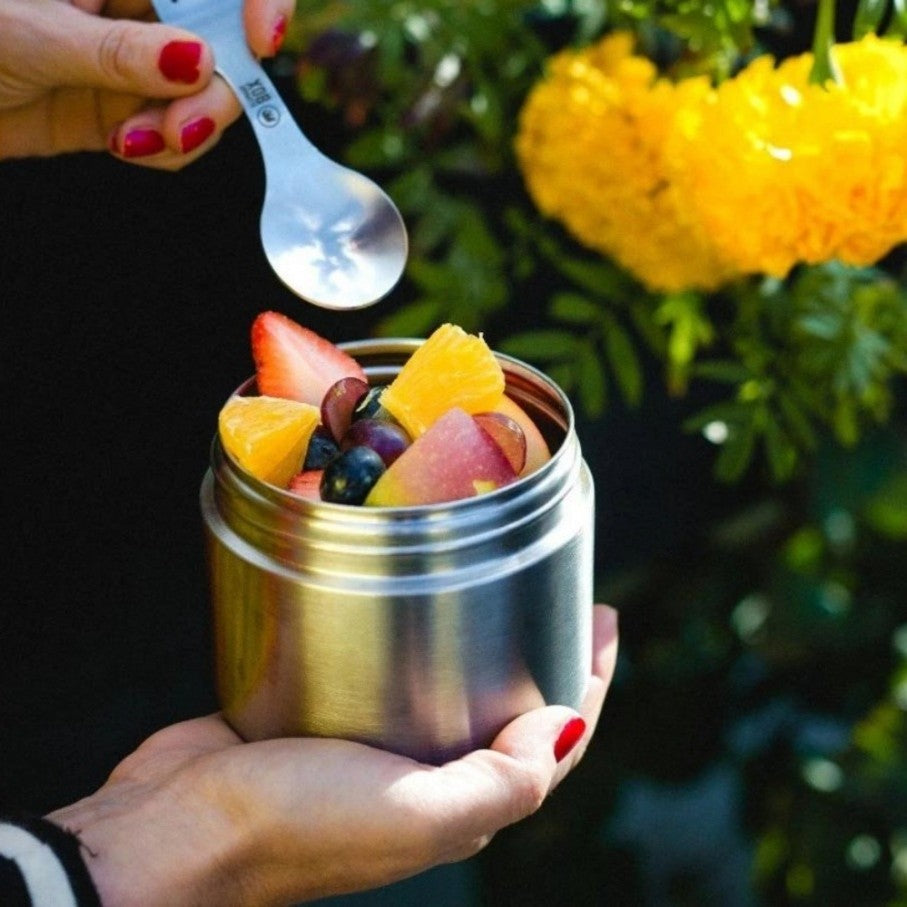 Stainless steel twist and lock food container being held in a hand with some fresh fruit and a spork digging in